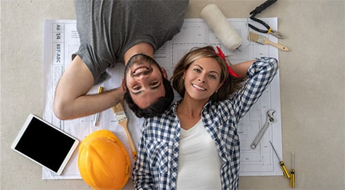 Couple laying down and looking up smiling at camera with paint roller and construction hat nearby